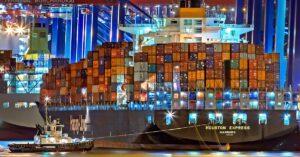 A brightly lit cargo ship at Hamburg harbor with stacked containers and a tugboat.